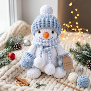 May include: A plush white crocheted snowman wearing a light blue ribbed hat with a pom-pom and a matching striped scarf. The snowman has a bright orange carrot nose, dark eyes, and light blue mittens. A brown tag reads Tiny Loop Studio.