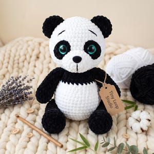 May include: A handcrafted crochet panda plush toy made of soft white and black yarn. The panda features large, glossy teal eyes, a small black nose, and a brown cardboard tag attached to its arm with twine that reads Tiny Loop Studio.