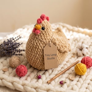 Peut inclure: Une poule en crochet beige avec une crête rose, un bec jaune et des yeux noirs. Une étiquette en papier brun attachée avec de la ficelle porte l'inscription Tiny Loop Studio. La poule est entourée de pelotes de laine, lavande et crochet.