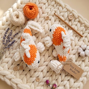 Könnte beinhalten: Zwei gehäkelte Koi-Fische aus weißem und orangefarbenem Garn mit schwarzen Augen und Flossen. Ein Fisch hat ein braunes Papieretikett mit der Aufschrift Tiny Loop Studio, das mit einer Schnur befestigt ist. Alles liegt auf einer groben Strickdecke.