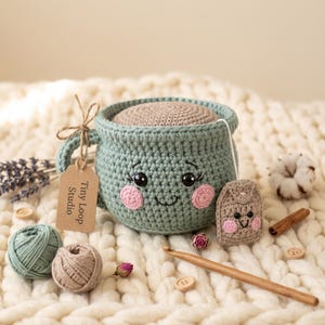 Könnte beinhalten: Eine salbeigrüne gehäkelte Tasse mit beigem Deckel, die ein lächelndes Gesicht mit rosa Wangen und schwarzen Augen zeigt. Ein kleiner beiger gehäkelter Teebeutel mit Gesicht ist an einer weißen Schnur befestigt. Ein braunes Etikett Tiny Loop Studio hängt am Henkel.