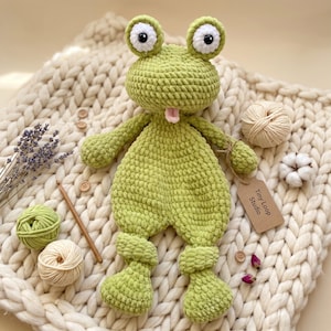Peut inclure: Une peluche grenouille crochetée vert clair avec de grands yeux blancs et noirs et une petite langue rose. La grenouille a des bras et des jambes noués et une étiquette en papier brun attachée au bras portant l'inscription Tiny Loop Studio.