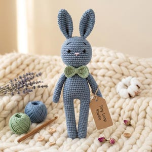 May include: A blue crocheted rabbit doll with long ears, black bead eyes, and a light green bow tie. The rabbit has a brown cardboard tag attached to its arm that reads Tiny Loop Studio in black text, featuring a simple stitched construction.