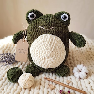 Op de afbeelding: Een donkergroene gehaakte kikkerknuffel met een grote crèmekleurige buik, zwart-witte plastic ogen en een kleine geborduurde mond. Een bruin papieren label aan de arm bevat de zwarte tekst Tiny Loop Studio, vergezeld door garenbollen.