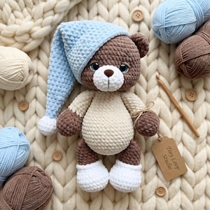 Könnte beinhalten: Ein gehäkelter Teddybär mit dunkelbraunem Fell, einem cremefarbenen Bauch und weißen Füßen. Der Bär trägt eine hellblaue, texturierte Schlafmütze mit einem weißen Bommel. Ein braunes Papieretikett am Arm trägt die Aufschrift Tiny Loop Studio.