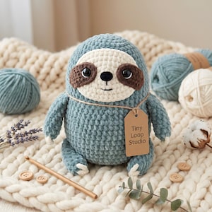 Può includere: Un peluche a forma di bradipo lavorato all'uncinetto in blu polvere con viso color crema e macchie marrone scuro intorno agli occhi. Presenta occhi di sicurezza neri, naso e bocca ricamati e un cartellino in cartone beige con scritto Tiny Loop Studio.
