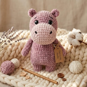 May include: A plush, mauve-colored crocheted hippopotamus toy with large black eyes and a lighter-toned snout. A brown paper tag attached to its arm reads Tiny Loop Studio. The toy is crafted from soft, textured yarn and features a rounded body.