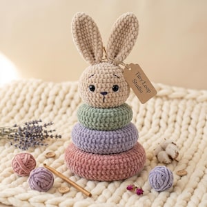 Op de afbeelding: Een gehaakt stapelspeelgoed met een beige konijnenkop als bovenkant, zwarte ogen en een geborduurde neus, rustend op drie zachte ringen in saliegroen, lavendel en oudroze. Een bruin papieren label aan het oor vermeldt Tiny Loop Studio.