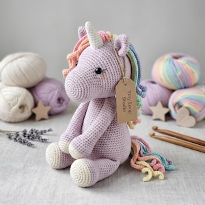 May include: A lavender crochet unicorn plush toy with a white horn, a multi-colored pastel mane, and a matching tail. A brown cardboard tag attached to the ear features the text Tiny Loop Studio. The unicorn has black eyes and pink cheeks.