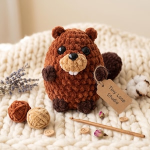 May include: A brown crocheted beaver plush toy with black plastic eyes, a tan snout, and two white teeth. A brown paper tag attached to its arm reads Tiny Loop Studio. The plush is surrounded by yarn balls, a wooden crochet hook, and dried flowers.