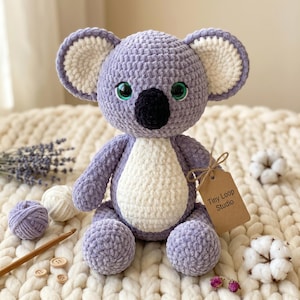 Può includere: Un peluche koala all'uncinetto realizzato in morbido filato viola chiaro con pancia e interno orecchie bianchi. Presenta grandi occhi verdi e un naso nero. Un cartellino di carta marrone attaccato al braccio riporta il testo Tiny Loop Studio.