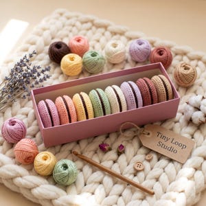 May include: A pink rectangular box containing eight crocheted macaron-shaped ornaments in various pastel colors, each with a dark brown center. The set includes small yarn balls, a wooden crochet hook, two buttons, and a tag labeled Tiny Loop Studio.