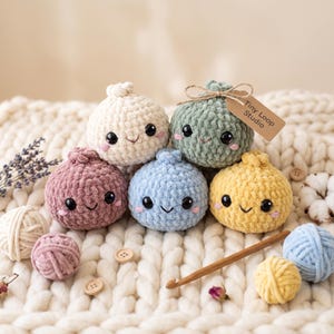 Puede incluir: Cinco criaturas redondas de peluche tejidas a ganchillo en color crema, verde salvia, rosa viejo, azul claro y amarillo. Cada una tiene ojos de seguridad negros, una sonrisa bordada y mejillas rosadas. Una etiqueta marrón dice Tiny Loop Studio.