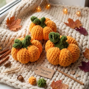 Puede incluir: Tres calabazas de ganchillo naranjas con tallos y hojas de color verde oscuro. Se adjunta una etiqueta de papel color canela con el texto Tiny Loop Studio. Las calabazas están sobre una manta de punto color crema.
