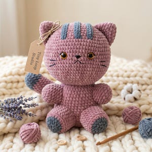 May include: A mauve crochet cat plush toy with dark gray stripes on its head and dark gray paws. The cat has brown plastic eyes, embroidered whiskers, and a brown paper tag attached to its ear with twine that reads Tiny Loop Studio.