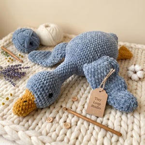 May include: A blue crocheted duck plush toy with a yellow beak and feet, featuring a brown cardboard tag labeled Tiny Loop Studio. The plush is surrounded by blue and white yarn balls, a wooden crochet hook, and dried lavender sprigs.