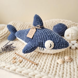 May include: A plush shark toy made of dark blue and white yarn with a soft, textured finish. It features a small beige tag tied with twine that reads Tiny Loop Studio. The shark has embroidered black gill slits and a white eye with a black pupil.