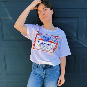 May include: A white t-shirt with a blue and red Budweiser logo printed on the front. The logo features the text "Budweiser" and "Genuine" on either side of the logo.