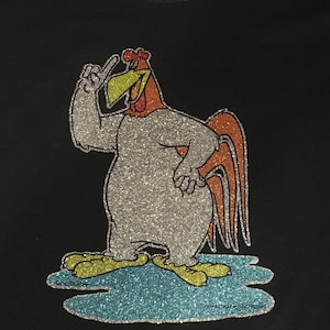May include: A black t-shirt featuring a silver glitter design of a cartoon rooster with a yellow beak and red comb. The rooster is standing on a blue glitter puddle.
