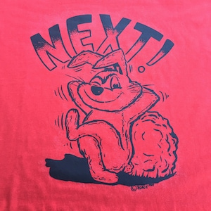May include: Red t-shirt with a black cartoon rabbit wearing a hat and the word "NEXT!" printed on it. The rabbit is sitting on its haunches with its arms crossed. The text "ROACH PIX" is printed in the bottom right corner.