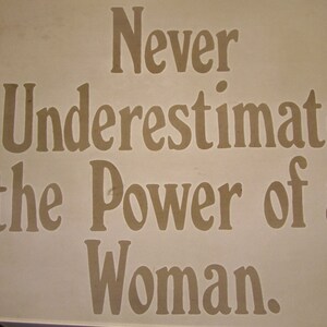 Puede incluir: Un cartel blanco con el texto "Never underestimate the power of a woman." en letras marrones.