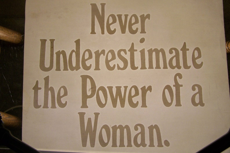 Puede incluir: Un cartel blanco con el texto "Never underestimate the power of a woman." en letras marrones.