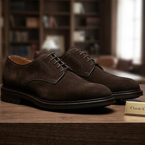 Peut inclure: Une paire de chaussures derby en daim marron foncé avec des lacets assortis et une semelle sombre. Une petite plaque rectangulaire dorée est placée devant les chaussures, portant le texte gravé 'Classic Clement Derby' en police serif.