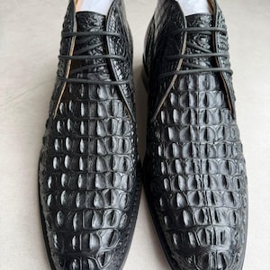 May include: A pair of black ankle boots featuring a textured crocodile skin pattern. These boots have a rounded toe, a lace-up closure with thin black laces, and white paper inserts visible inside the top opening of each shoe.