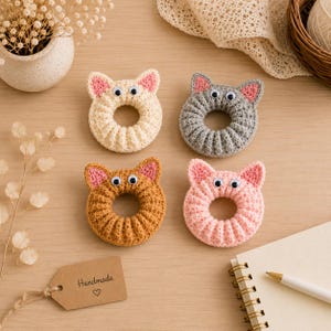 Könnte beinhalten: Vier kreisförmige Häkelringe in Katzenform in Creme, Grau, Braun und Pink. Jeder Ring hat zwei spitze Ohren mit rosa Innendetails und zwei Wackelaugen aus Kunststoff. Ein beigefarbenes Pappschild mit dem Wort Handmade und einem Herzsymbol.