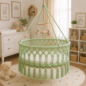 May include: A round, light green macrame hanging cradle featuring a complex knotted pattern and decorative fringe along the bottom edge. The cradle is suspended by thick cords and includes a central mobile with small plush animal figures.