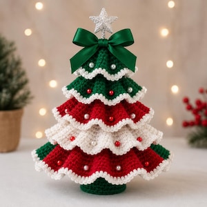 Puede incluir: Una decoración de árbol de Navidad tejida a ganchillo con volantes escalonados en colores alternos verde oscuro, rojo y crema. Los bordes están ribeteados en blanco y el árbol está adornado con pequeñas cuentas rojas y blancas, rematado con un lazo de satén verde y una estrella plateada.