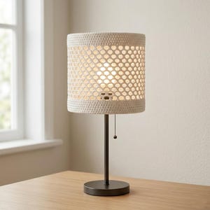 May include: A table lamp featuring a cylindrical lampshade made of off-white crocheted fabric with a repeating hexagonal pattern. The lamp has a slender dark brown metal stem and a circular base, complemented by a small pull chain switch.