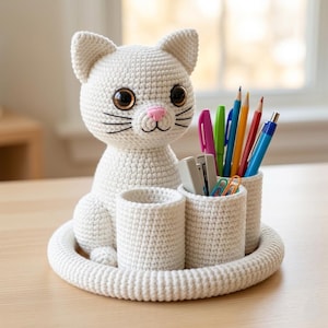 Puede incluir: Un organizador de escritorio de ganchillo blanco con forma de gato, que presenta una cabeza grande con ojos marrones, nariz rosa y bigotes negros. La base incluye tres compartimentos cilíndricos para bolígrafos, lápices y clips.
