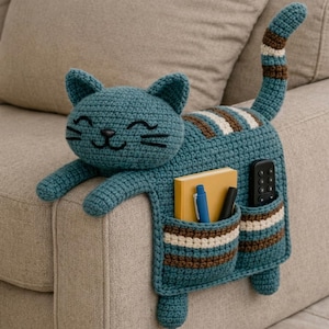 以下が含まれることがあります： 長い縞模様の尻尾、2つのフロントポケット、刺繍された顔を持つ、青緑色の猫型かぎ針編みアームレストオーガナイザー。ポケットには黄色いノート、青いペン、黒いマーカー、黒いリモコンが収納されています。