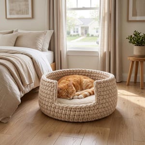 Op de afbeelding: Een rond, beige, grof gebreid huisdierenmandje met een dikke gevlochten rand en een zacht, crèmekleurig binnenkussen. Een oranje cyperse kat ligt opgerold te slapen in het mandje dat op een lichte houten vloer in een slaapkamer staat.