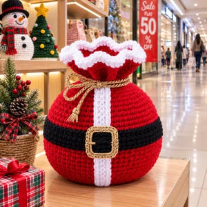 Pode incluir: Um saco de presente redondo em crochê vermelho com uma borda superior branca, um cinto preto com uma fivela dourada e uma faixa vertical branca. O saco é fechado no topo com um cordão trançado dourado e borlas.