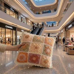 May include: A square throw cushion featuring a multi-coloured crochet granny square pattern in shades of peach, sage green, cream, and dusty rose. The cushion has a scalloped cream-coloured crochet border along all four edges of the square cushion.