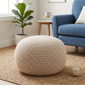 May include: A round, beige, crocheted pouf ottoman with a dense, textured knit pattern. The pouf is spherical in shape and features a consistent, neutral-colored yarn construction throughout its exterior, providing a firm and structured appearance.