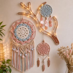 May include: A wall-mounted copper-colored metal branch structure with integrated fairy lights, featuring three circular dreamcatchers. The largest has a cream, blue, and pink crochet center with macrame fringe and beads. Two smaller dreamcatchers feature crochet, shell-like discs, and metal feather charms.