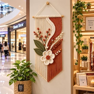 May include: A macrame wall hanging featuring a cream and rust-coloured cord design with a central white flower, green leaves, and red berry accents. The piece is suspended from a wooden dowel with a white rope hanger against a neutral wall.