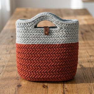 Puede incluir: Una cesta de ganchillo bicolor con una sección superior gris claro y una sección inferior color óxido, ambas tejidas en un denso patrón de espiga. La cesta incluye asas integradas y una pequeña lengüeta de cuero marrón con un broche metálico.