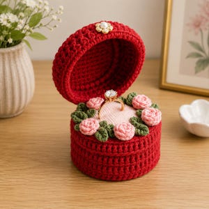 Könnte beinhalten: Eine runde, rote gehäkelte Ringbox mit offenem Deckel und einem weißen, perlenartigen Akzent. Das Innere ist mit einem weichen, hellrosa Kissen ausgekleidet, das von kleinen gehäkelten rosa Rosen und grünen Blättern umgeben ist und einen Goldring hält.