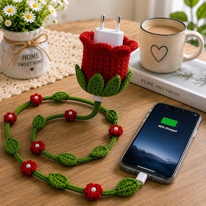 May include: A crochet cable protector featuring a red rose bud design that encases a white power adapter. The long green cord is decorated with small red crochet flowers and green leaves, connecting to a smartphone displaying 82% charge.