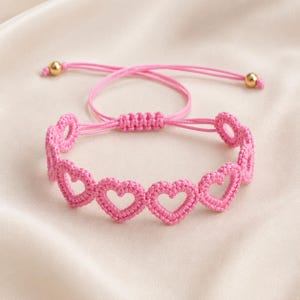 Peut inclure: Un bracelet rose composé d'une série de cœurs crochetés interconnectés. Le bracelet est doté d'un fermoir coulissant réglable avec deux fins cordons roses, chacun terminé par une petite perle en métal doré poli à l'extrémité.