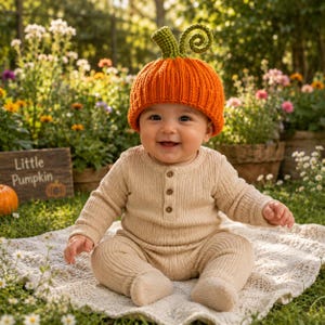 May include: A baby wearing a bright orange ribbed knit hat shaped like a pumpkin with a green stem and curly vine on top, paired with a long-sleeved beige ribbed onesie featuring three small buttons down the center of the chest area.