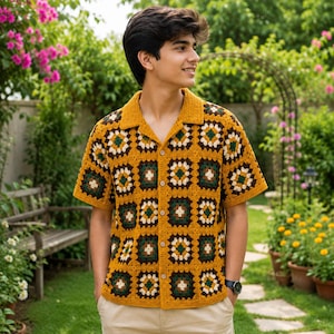 Puede incluir: Una camisa de manga corta con botones, confeccionada con cuadrados de ganchillo en tonos amarillo mostaza, verde oscuro y crema. La prenda presenta un cuello clásico, una tapeta frontal con botones y un patrón geométrico repetitivo.