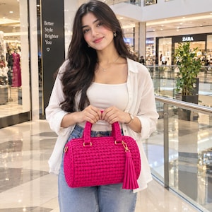 May include: A vibrant fuchsia cylindrical crochet handbag featuring two sturdy top handles, gold-toned metal hardware, and a long matching tassel charm attached to a side ring. The bag has a textured, woven yarn construction with a structured barrel shape.