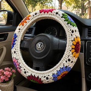 May include: A crochet steering wheel cover featuring a cream-colored base with a repeating pattern of multi-colored floral motifs in shades of red, orange, yellow, green, blue, and purple, fitted over a black steering wheel with a Volkswagen logo.