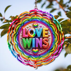 May include: A circular metal wind spinner featuring a rainbow-colored gradient pattern. The center displays the text LOVE WINS in bold, glittery, multi-colored 3D letters, accented by two rainbow-colored hearts and small star shapes on a white background.