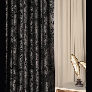 May include: A floor-length curtain panel featuring a black fabric with a textured, distressed silver metallic pattern. Beside it hangs a plain, vertical-pleated cream-colored curtain. Two gold-colored feather sculptures stand on a white base.